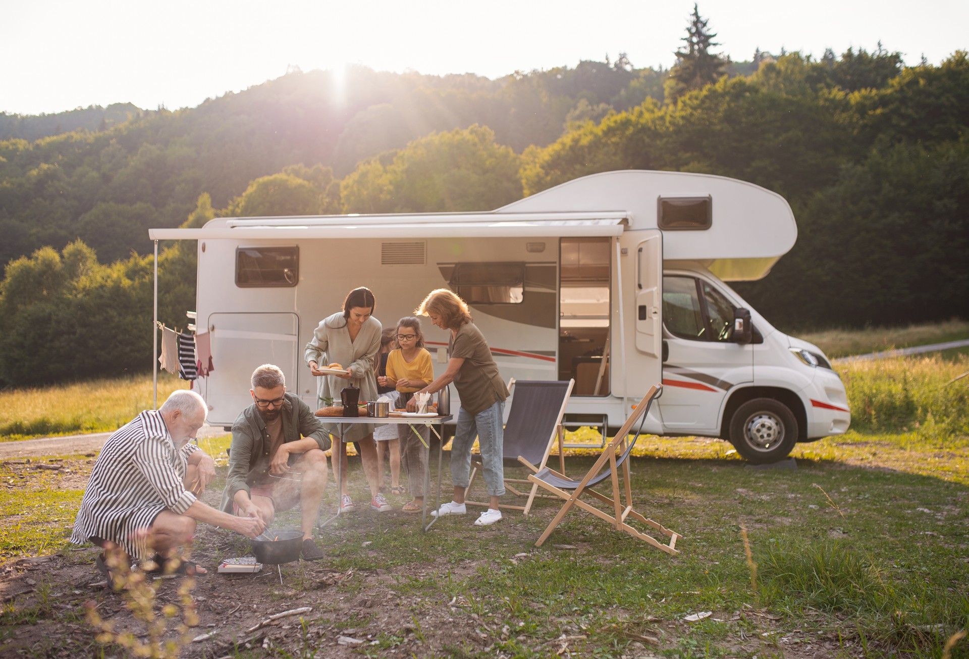 Multi-generation family sitting and eating outdoors by car, caravan holiday trip.